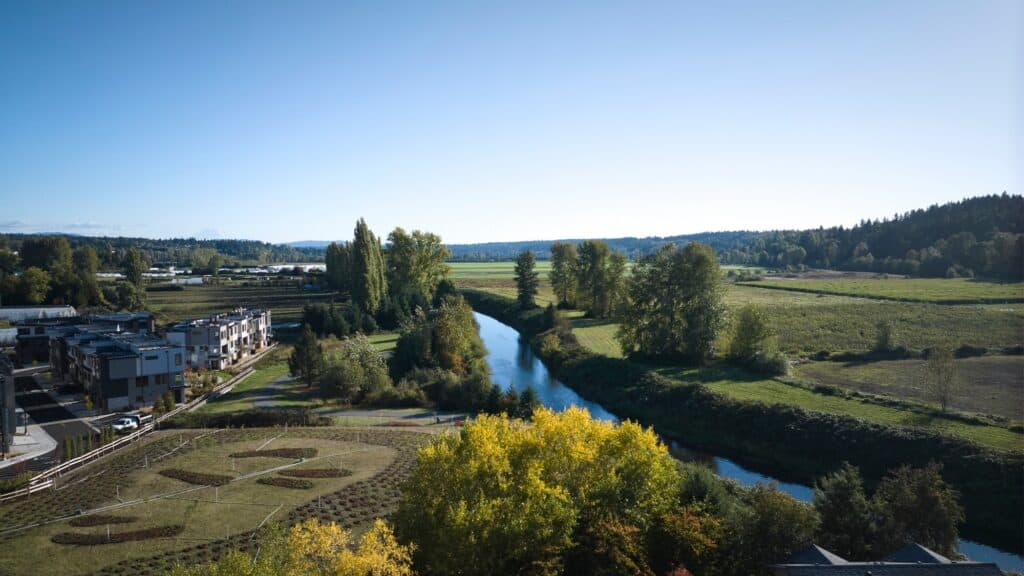 A river flows through a rural landscape with open fields, trees, and a nearby residential area under a clear blue sky.