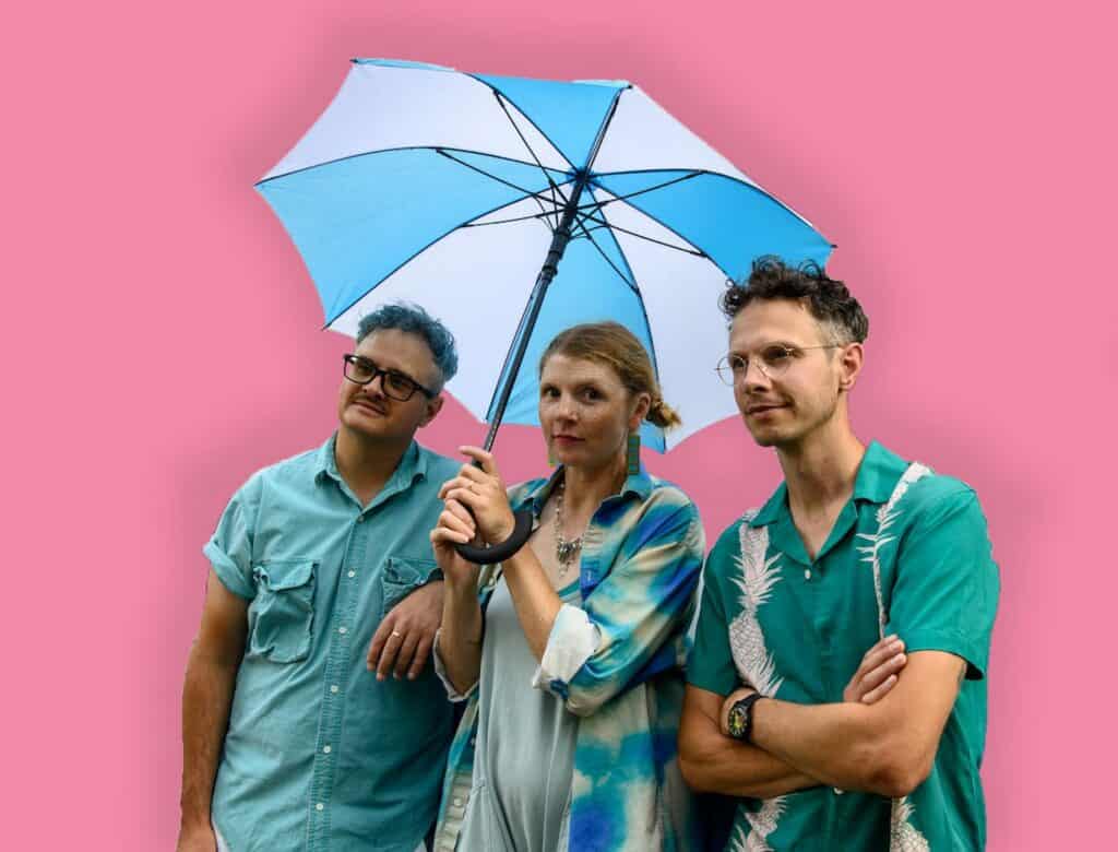 Three people stand in front of a pink background; the woman in the middle holds a blue and white umbrella, while the men on either side wear teal shirts.
