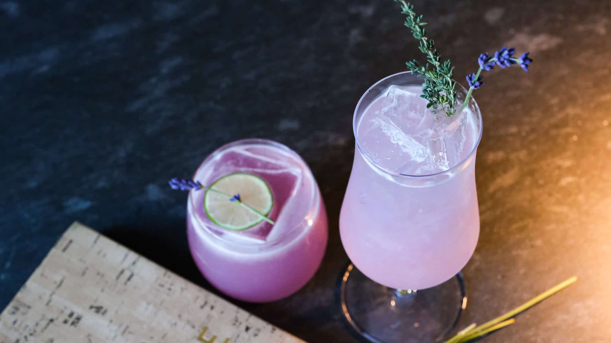 Two pink Covert Cocktails on a dark surface: one, the Lavender Last Call in a rocks glass with lime and lavender; the other in a stemmed glass with ice, thyme, and lavender. A Restaurant Roundup favorite for any occasion.