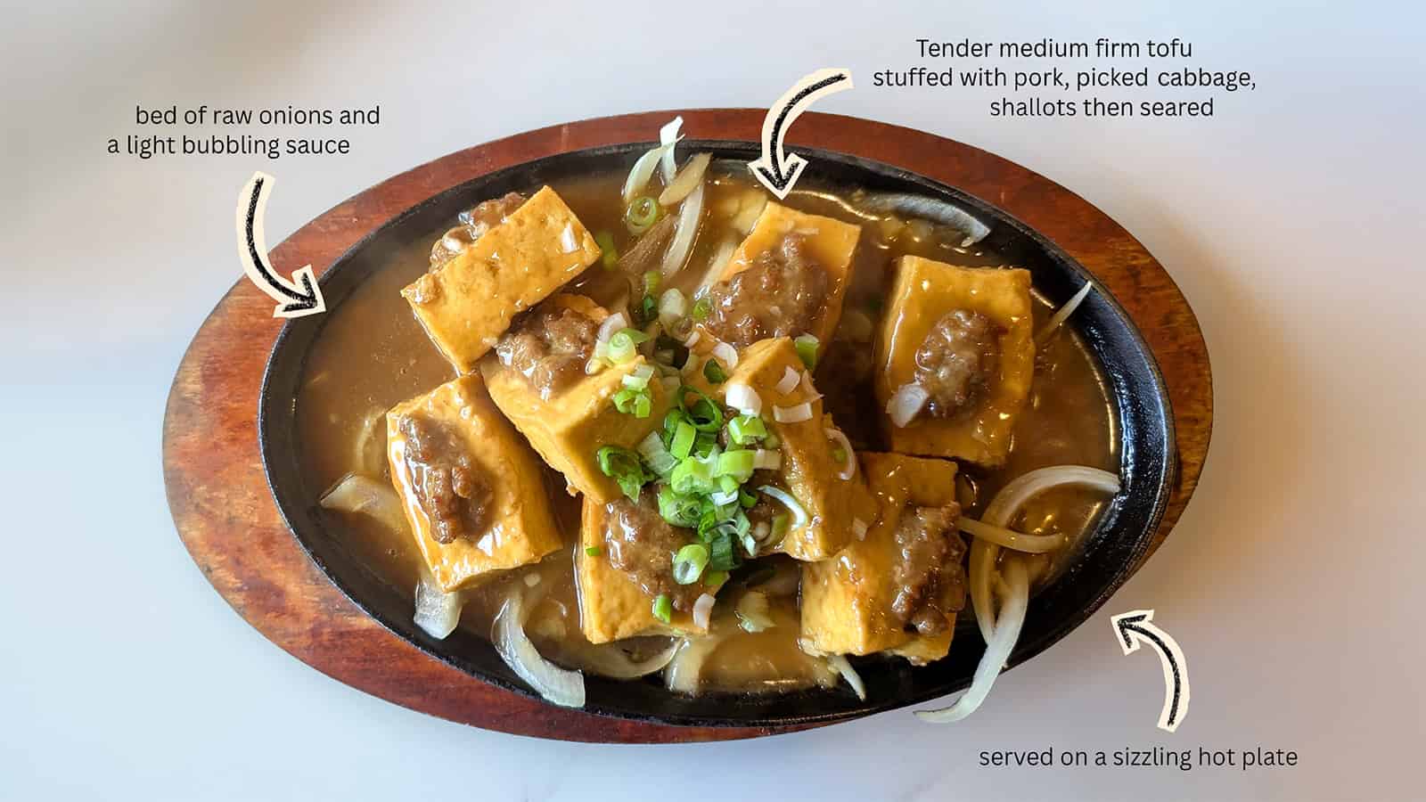 A sizzling hot plate of tofu stuffed with pork and vegetables, topped with sauce and green onions, served on raw onions in a light bubbling sauce.