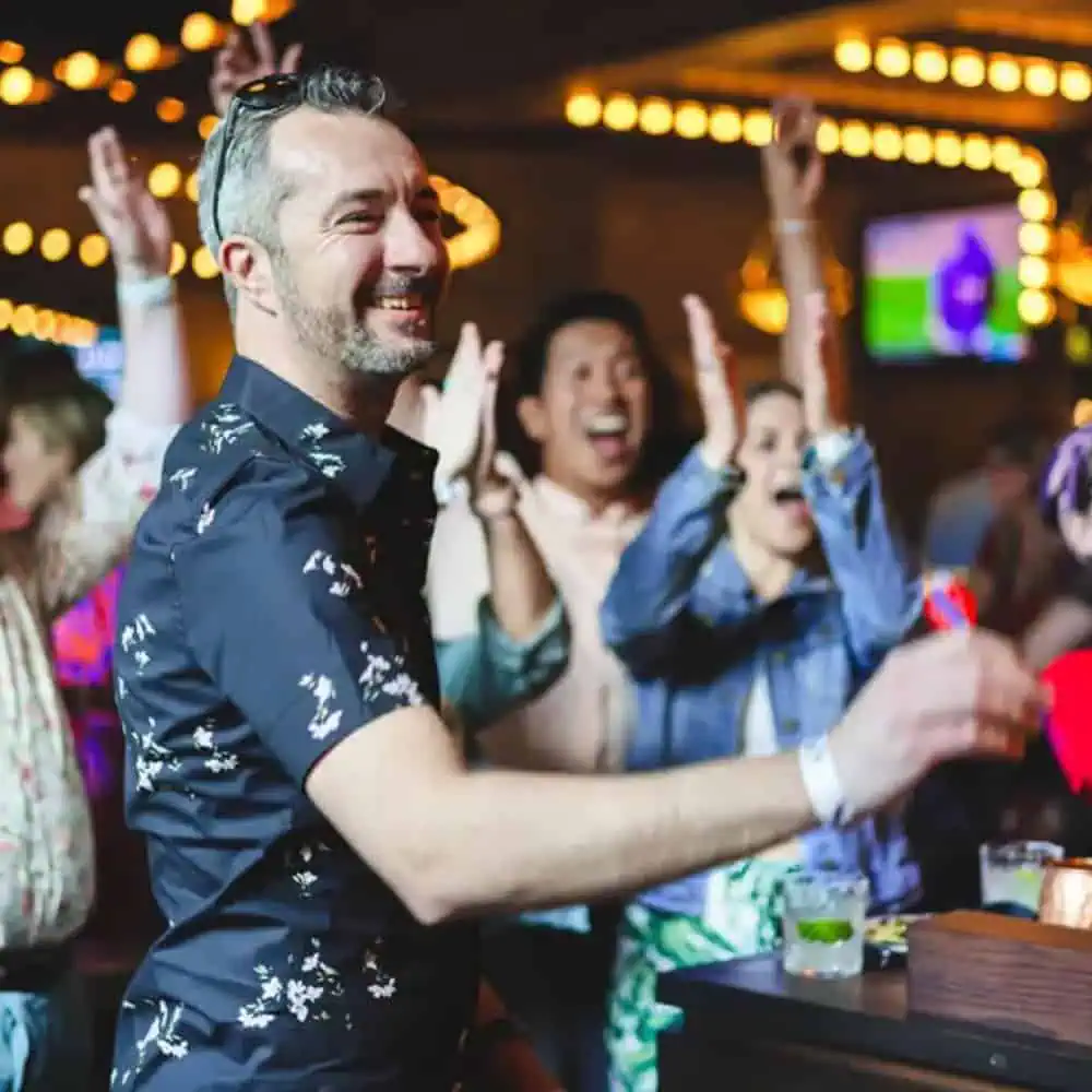 A man in a patterned shirt smiles and gestures in a lively bar, surrounded by people cheering with drinks—perfect for a Father’s Day celebration or inspiration for your next Gift Guide.
