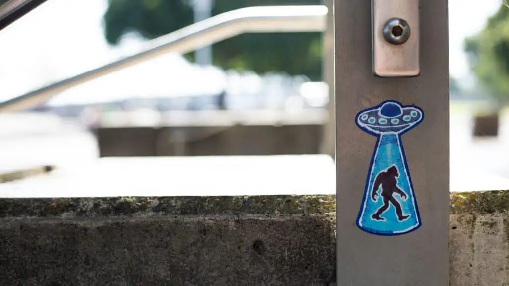 A sticker shows a UFO beaming up a silhouette of Bigfoot, placed on a metal rail near outdoor concrete steps.
