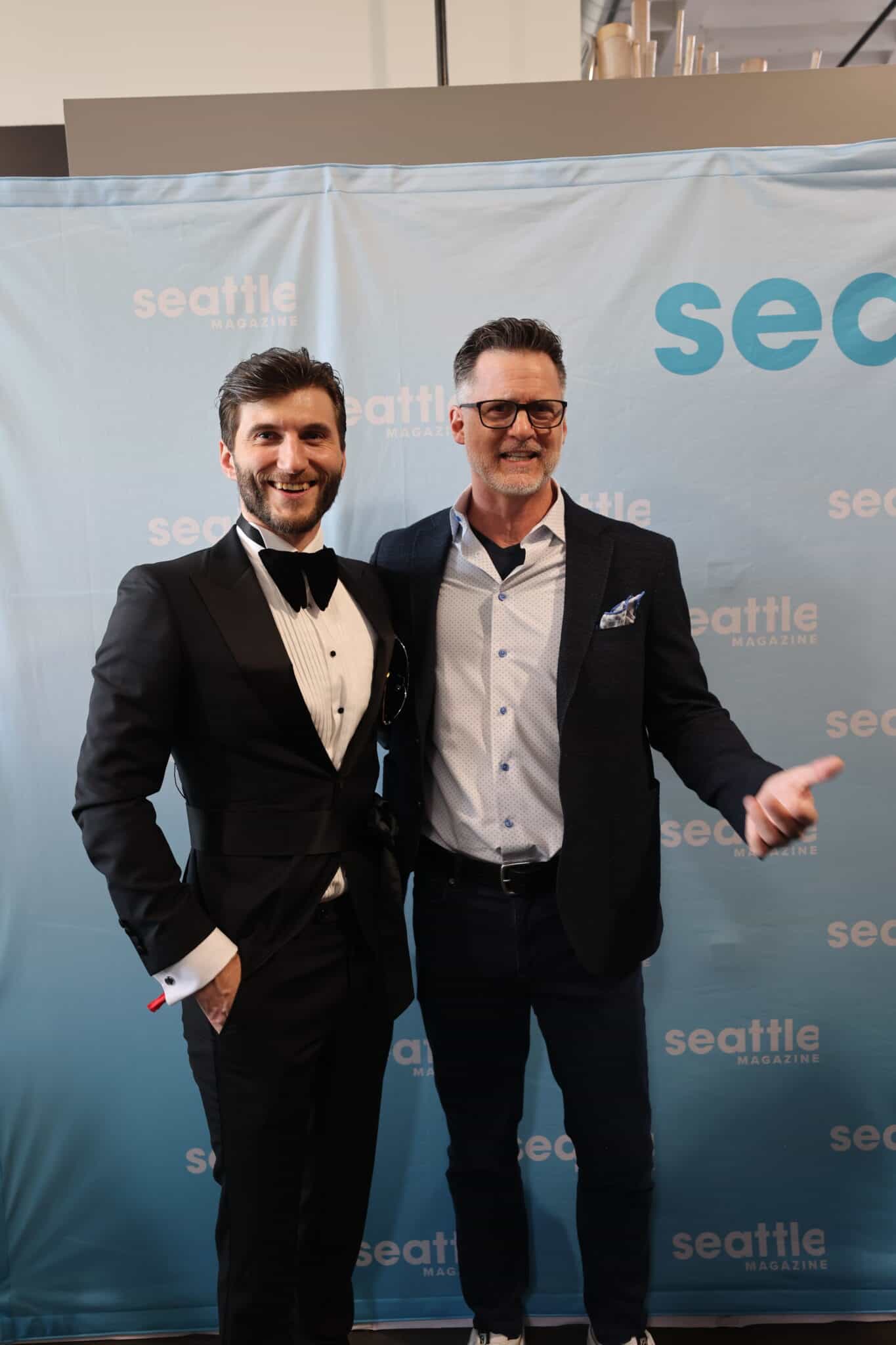 Two men pose and smile in front of a Seattle Magazine step-and-repeat banner; one wears a tuxedo, the other a blazer and dress shirt.