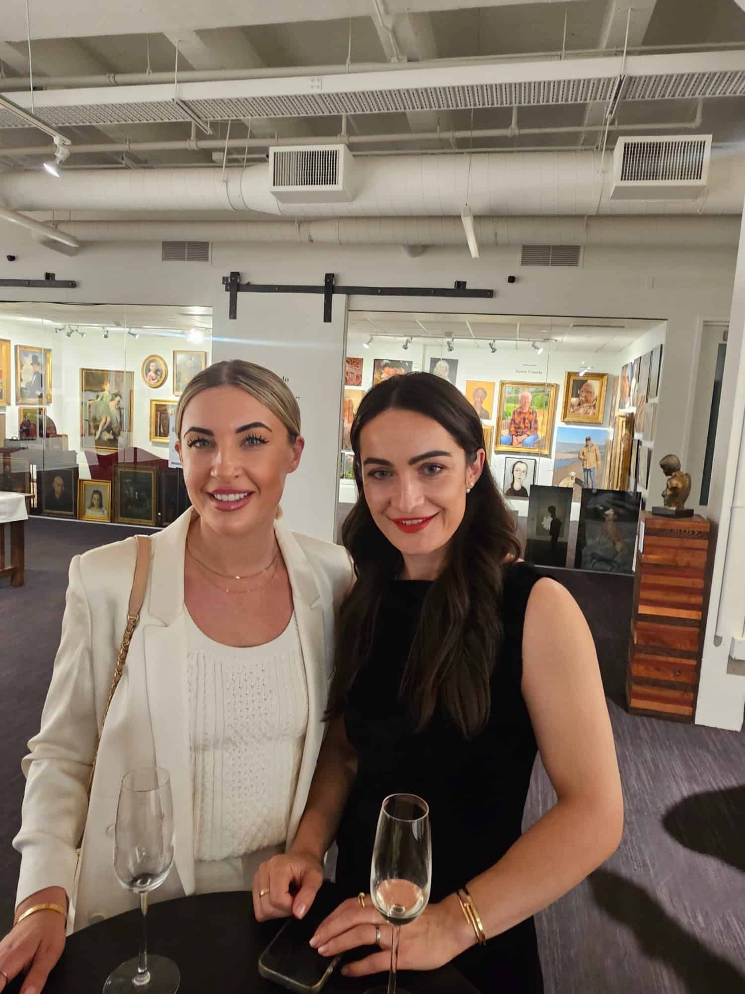 Two women stand together at a table in an art gallery, each holding an empty champagne glass, with paintings and sculptures visible in the background.