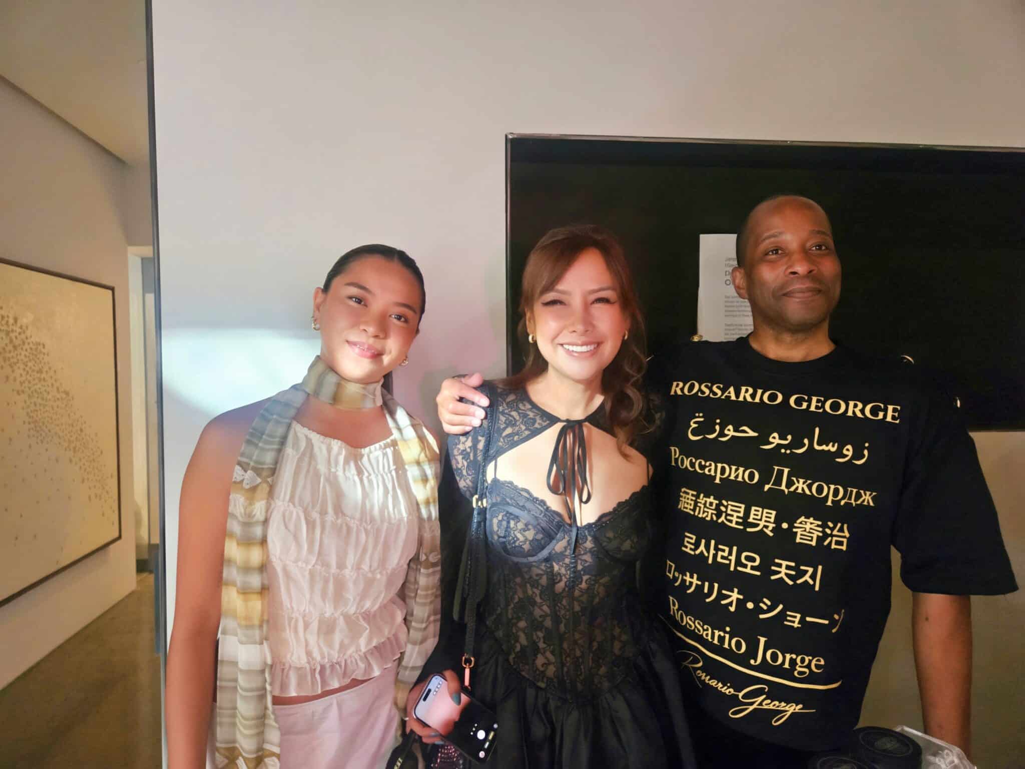 Three people stand indoors, posing and smiling for the camera. The person on the right wears a black printed shirt, and the person in the center wears a lacy black top.
