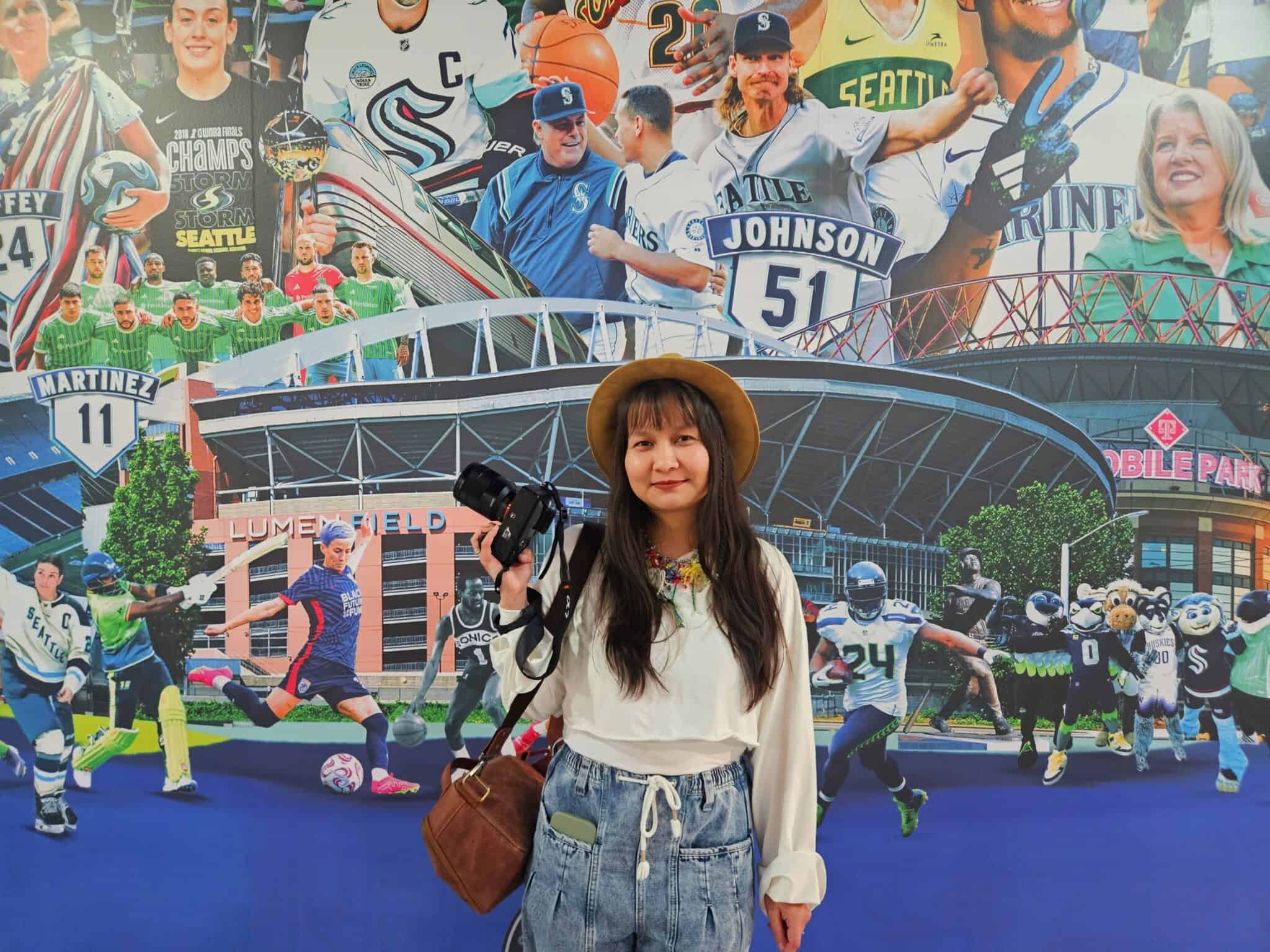A woman holding a camera stands in front of a mural featuring Seattle sports teams, athletes, and stadiums.