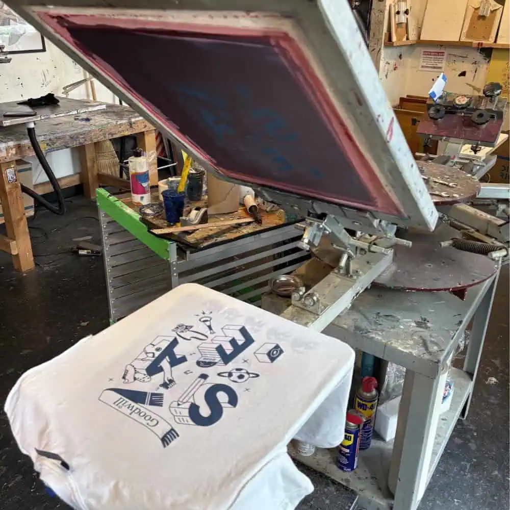 A screen printing machine with a partially printed white t-shirt displaying blue graphics and text in a workshop setting.