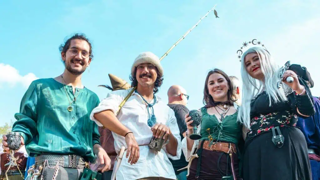 Four people in colorful medieval or fantasy costumes stand outdoors, smiling and holding cups, with a bright sky in the background.