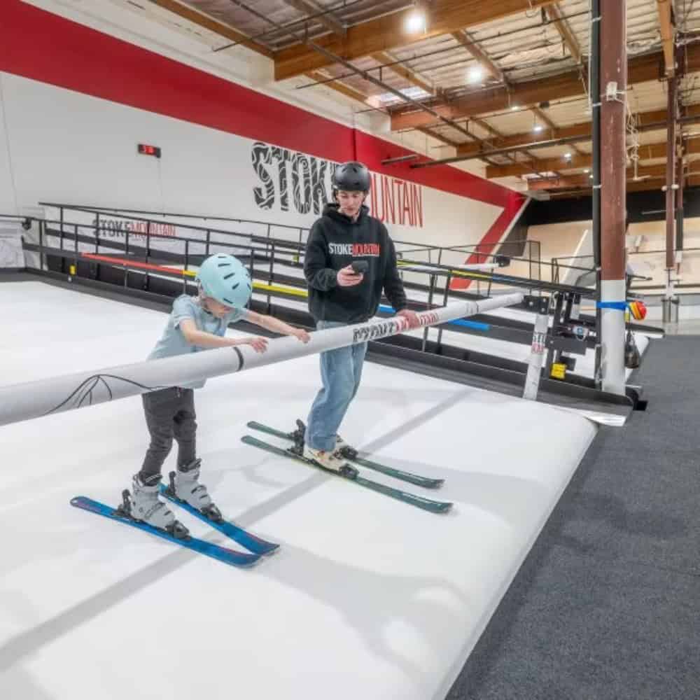 A child wearing a helmet practices skiing on an indoor ski slope while an adult beside them observes and uses a smartphone.