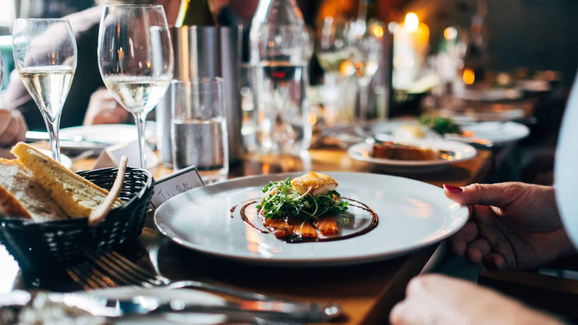 A plated gourmet dish with greens and sauce is placed on a wooden table set for dining, with wine glasses, bread, and candles visible.