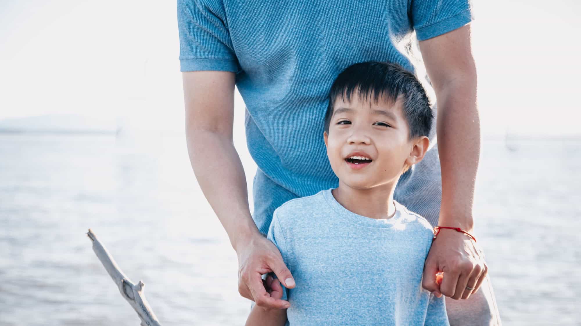 An adult stands behind a smiling boy by the water, both wearing blue shirts. The adult's hands rest gently on the boy's shoulders.