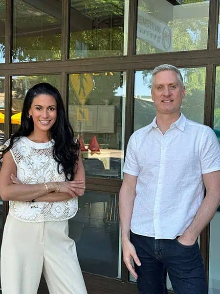 A woman and a man stand side by side in front of a glass building at Terminal One, both wearing light-colored, casual clothing and smiling at the camera.