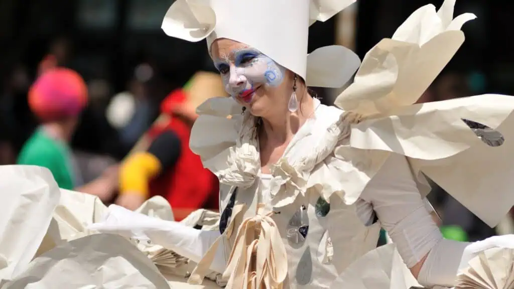 Person in a whimsical white costume with paper-like details and blue face paint, participates in an outdoor event or parade.