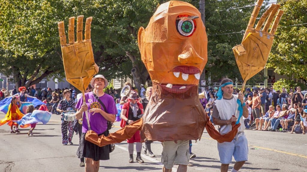 People parade down a street carrying and wearing a large, whimsical puppet of a one-eyed creature with big hands, while a crowd watches from the sidewalk.