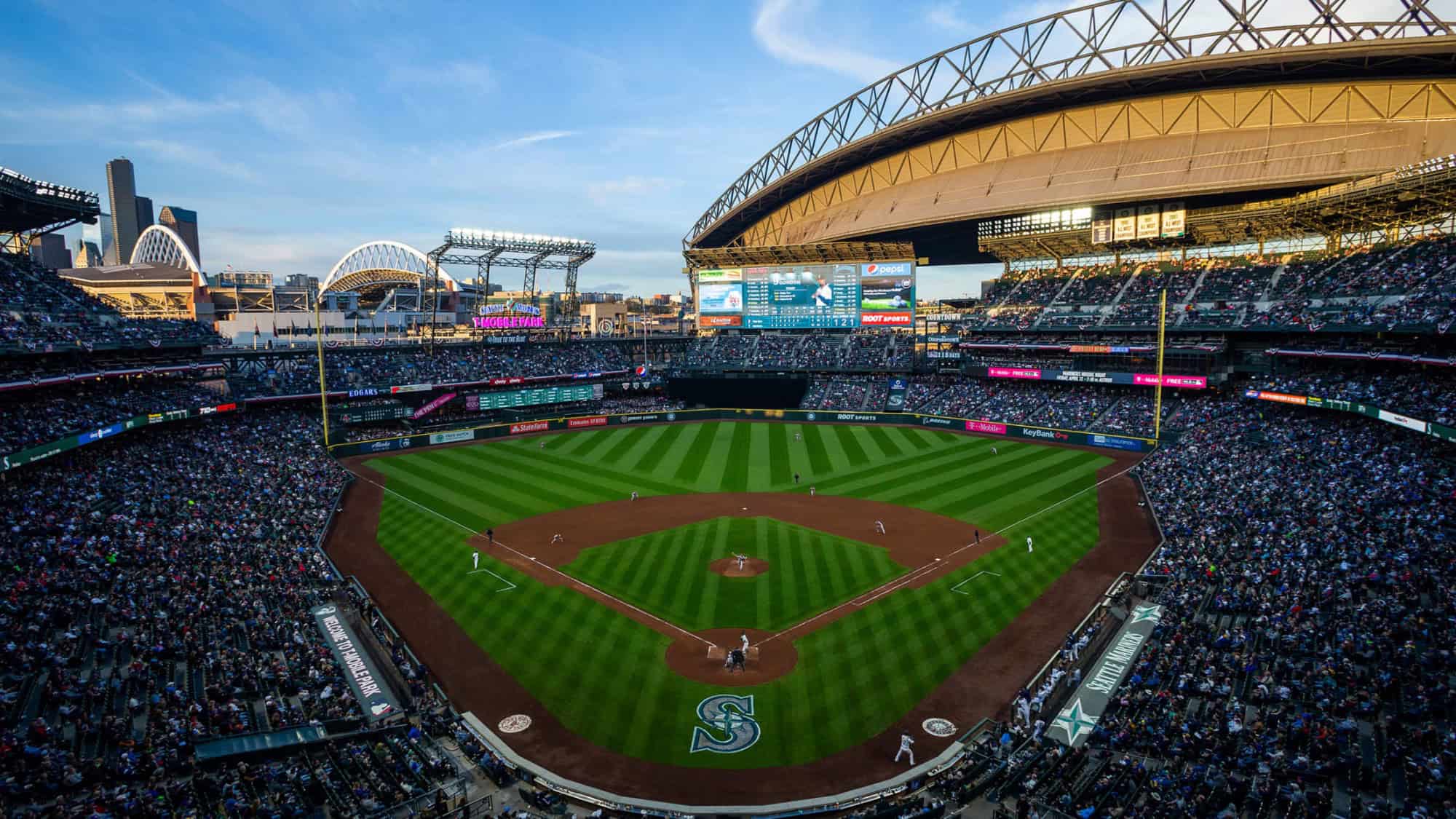 A packed baseball stadium viewed from behind home plate, with players on the field and a retractable roof partially open under a blue sky.