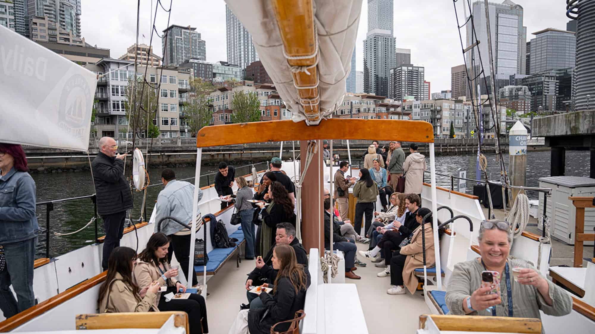 People sit and stand on a sailboat docked by a city waterfront, eating, talking, and taking photos; tall buildings and cloudy skies are visible in the background—a lively scene that captures the vibrant spirit of PNW vendors often showcased at events like the Northwest Event Show.