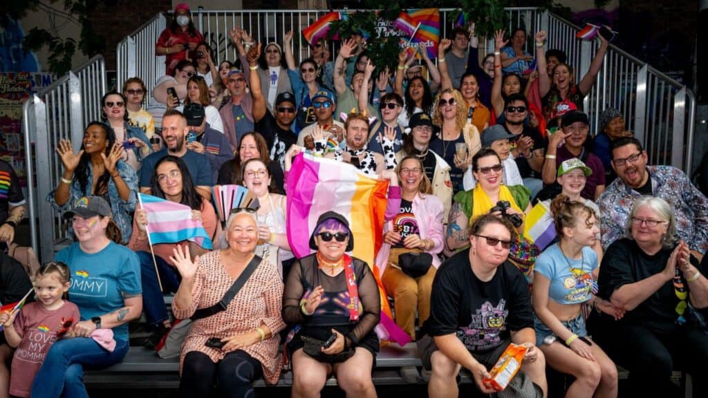 A diverse group of people sitting on bleachers, waving and holding LGBTQ+ pride flags at an outdoor event.