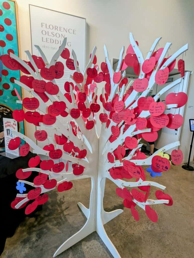 A white tree-shaped display holds numerous red paper apples, each with handwritten messages, set in an indoor exhibit area.