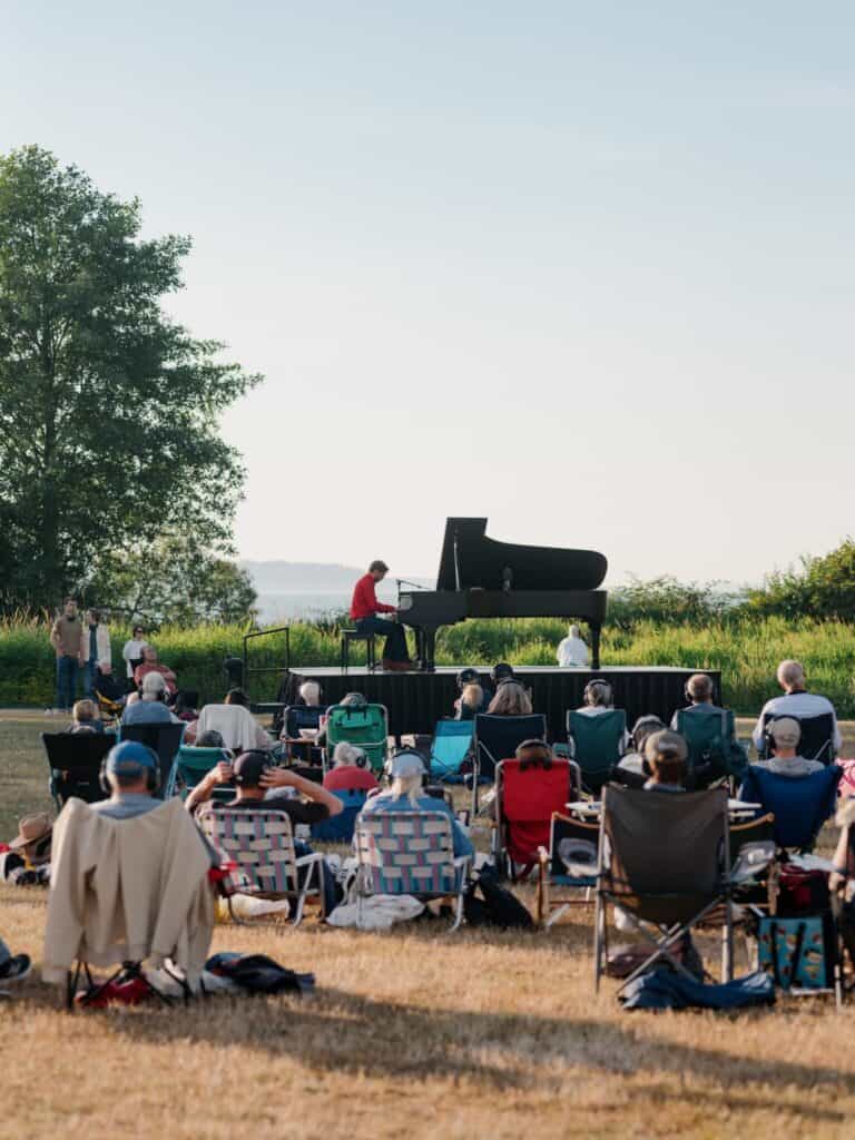 A pianist performs on a grand piano outdoors as an audience sits in lawn chairs on a grassy field under a clear sky.