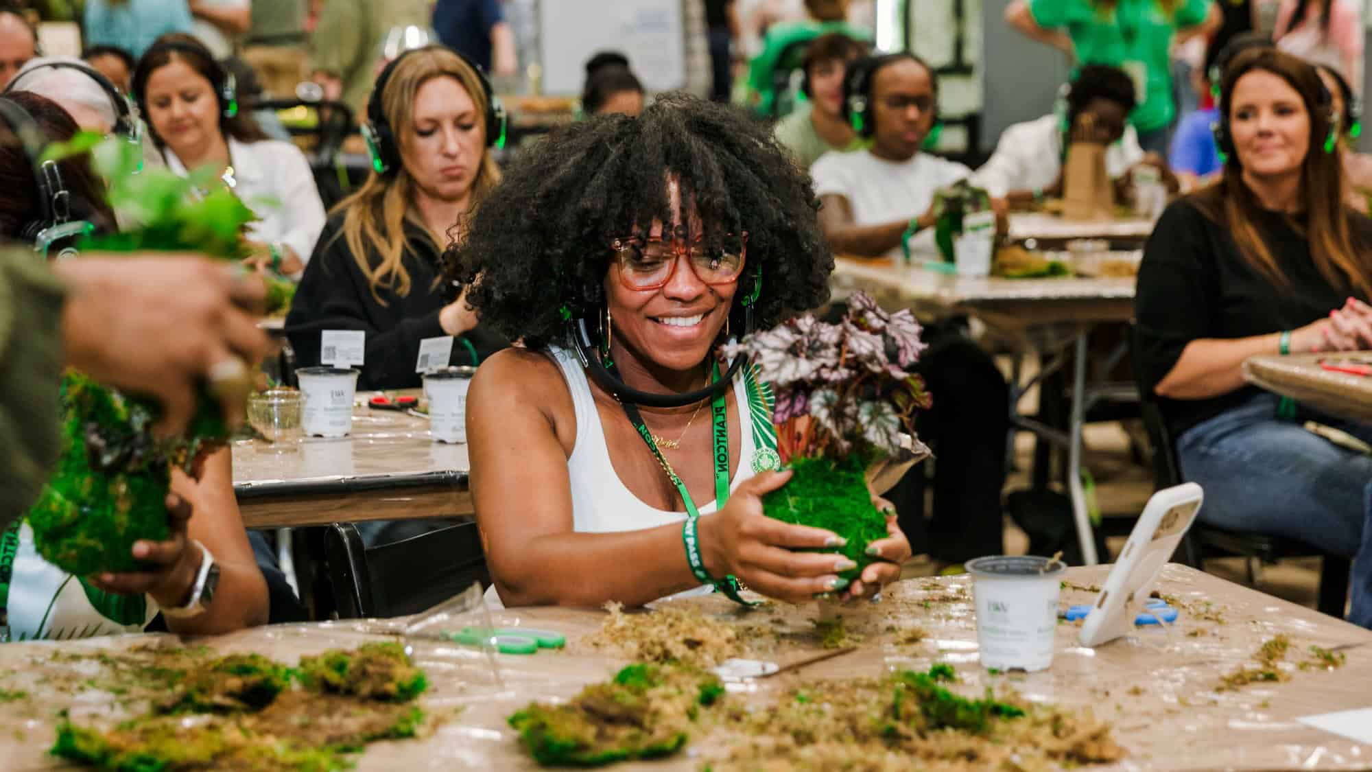 A woman smiles while making a moss plant arrangement at a table, surrounded by other people participating in the same group activity.