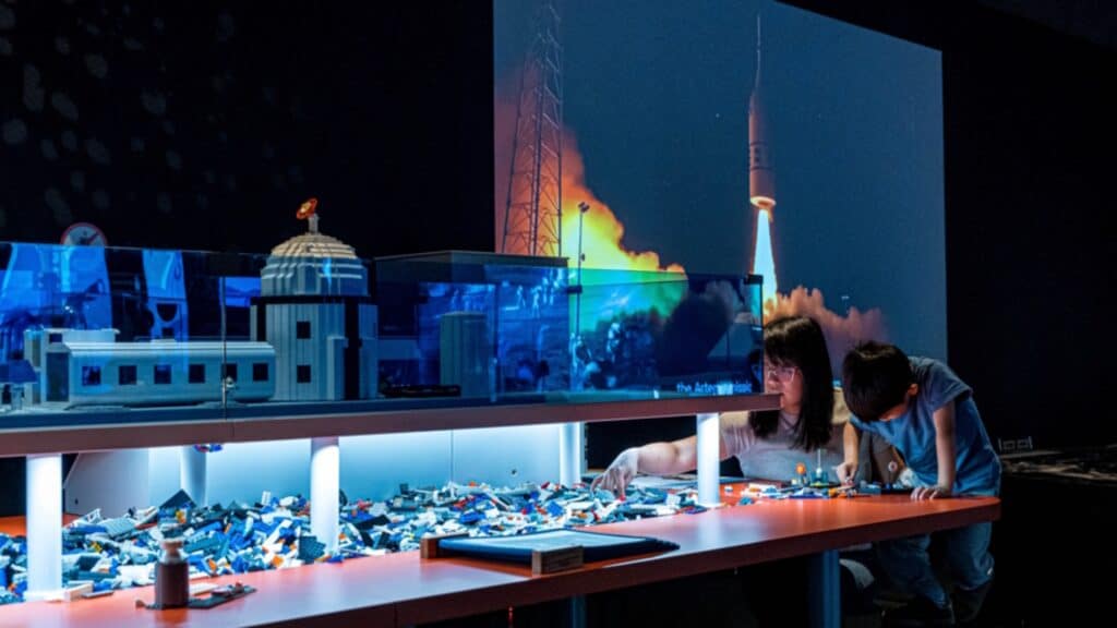 A woman and child build with LEGO bricks at a table, enjoying stress-free activities together, with a rocket launch projected on the wall and model buildings displayed behind them—perfect inspiration for Seattle family outings.