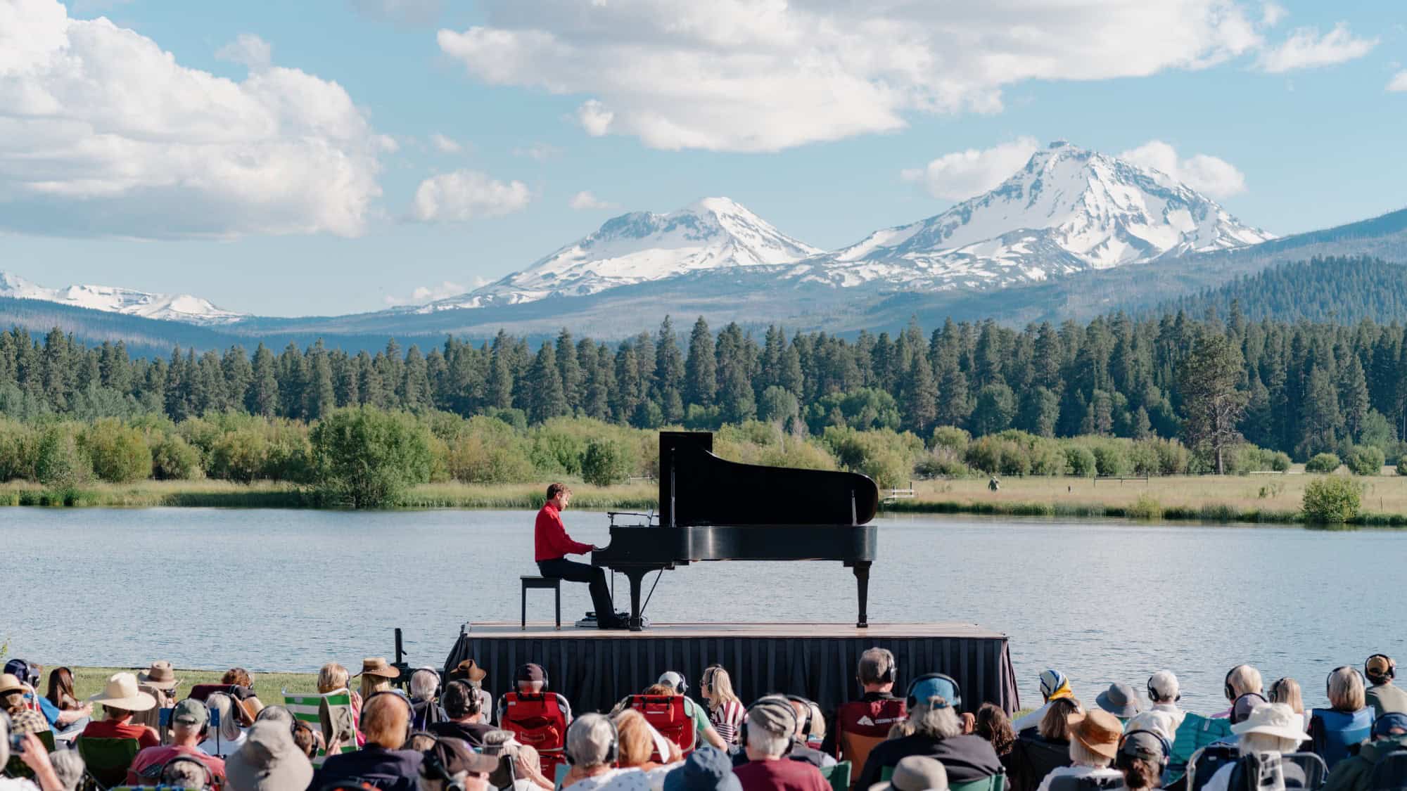 A pianist in a red shirt performs on an outdoor stage by a lake with mountains and pine trees in the background as an audience watches.
