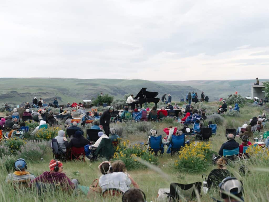 A crowd sits on lawn chairs and blankets listening to a pianist performing outdoors in a grassy, hilly landscape under a cloudy sky.