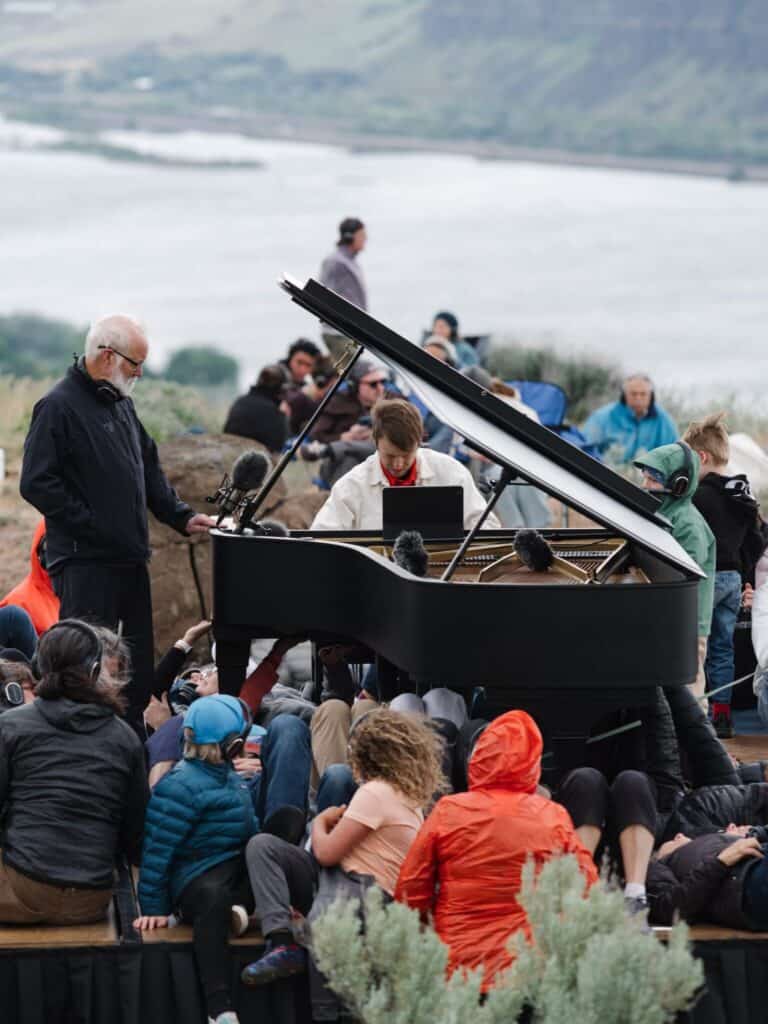 A group of people sit outdoors near a grand piano, where a person is playing music as others watch by a river with hills in the background.