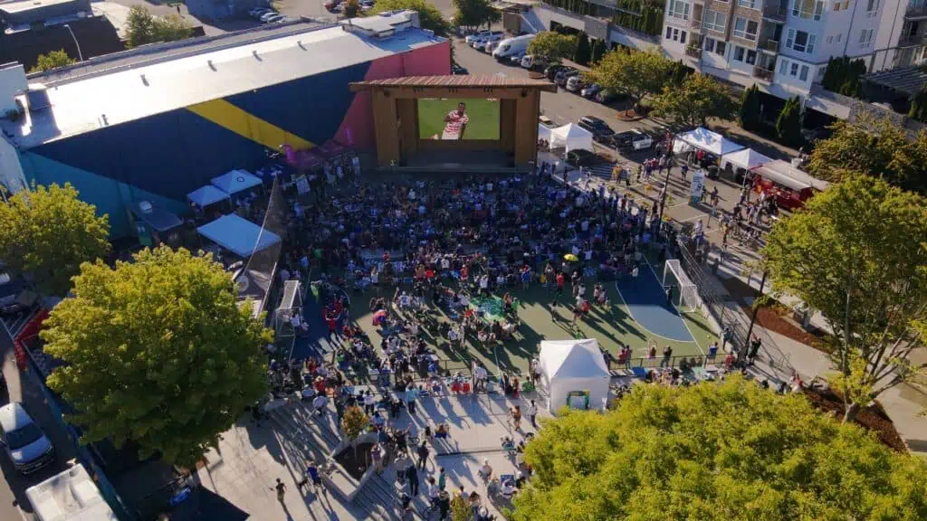 A large crowd gathers in an outdoor plaza during the day, watching a big screen near a colorful building, with trees, tents, and nearby parked cars visible.