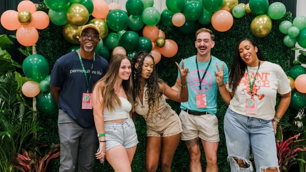 Five people pose in front of a green and orange balloon backdrop, smiling and making playful gestures, wearing event badges and casual summer clothes.