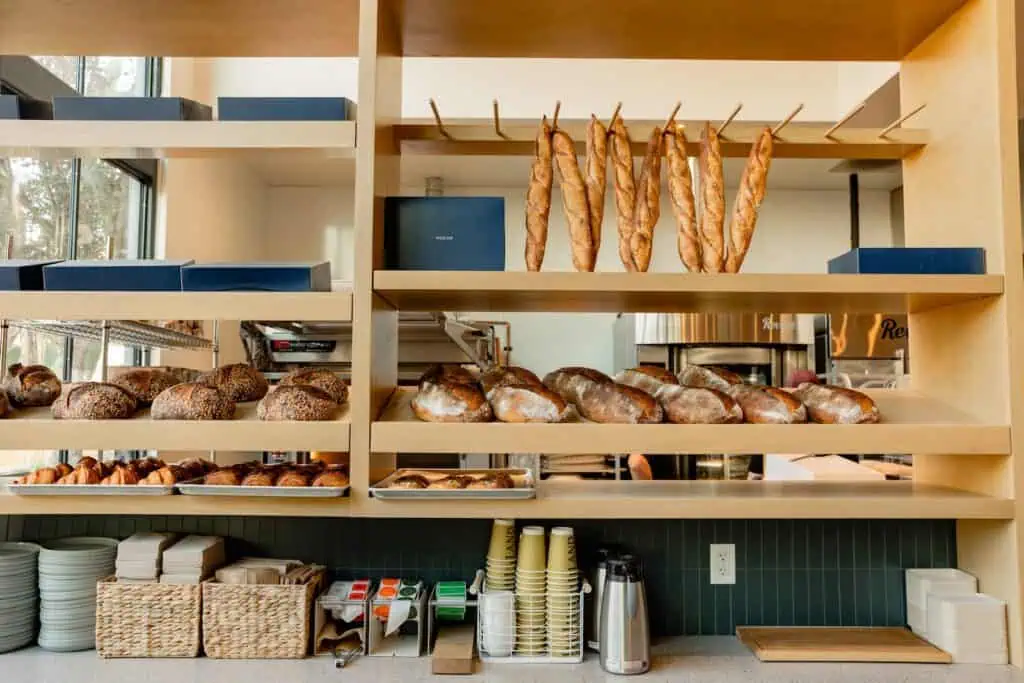 Shelves in a Carlsbad bakery display baguettes, round loaves, and pastries. Below are stacks of plates, cups, napkins, and coffee supplies on a counter—perfect for a relaxing weekend treat.