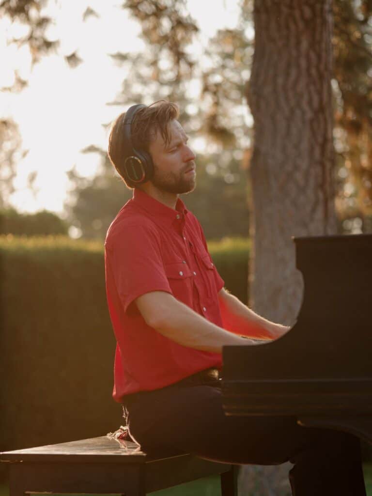 A man in a red shirt and headphones plays a grand piano outdoors near a tree, with sunlight filtering through the background.