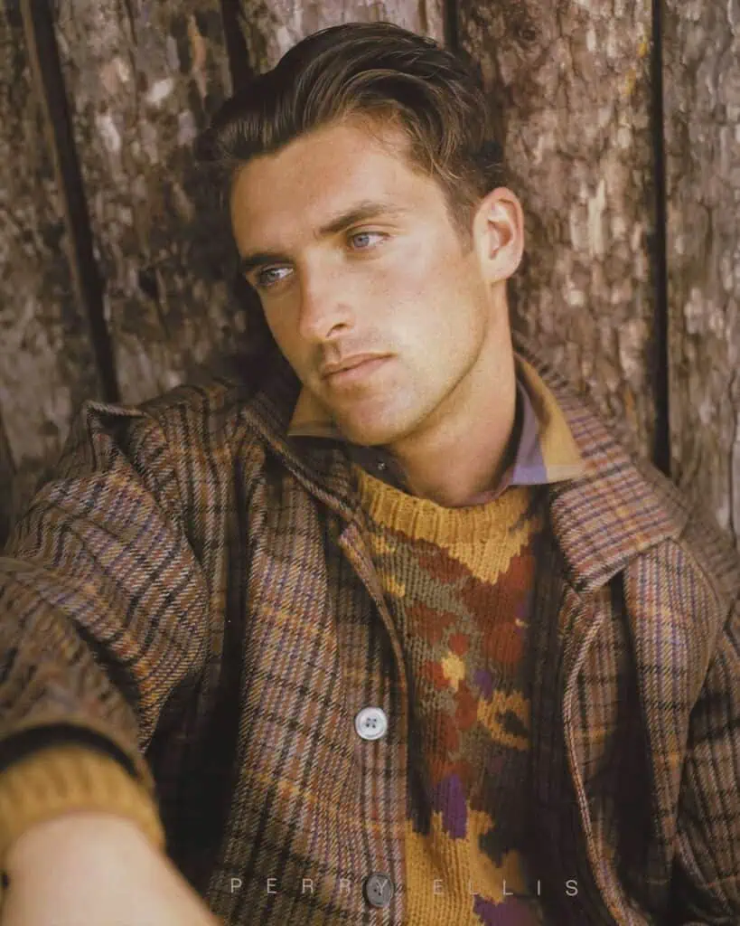 A man in a patterned brown jacket and sweater, embodying fashion and culture, sits before a large tree trunk, looking to the side with a contemplative expression.