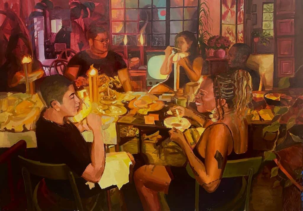A group of six people sit around a table with food and lit candles, sharing a meal in a warmly lit, colorful room filled with plants and art.
