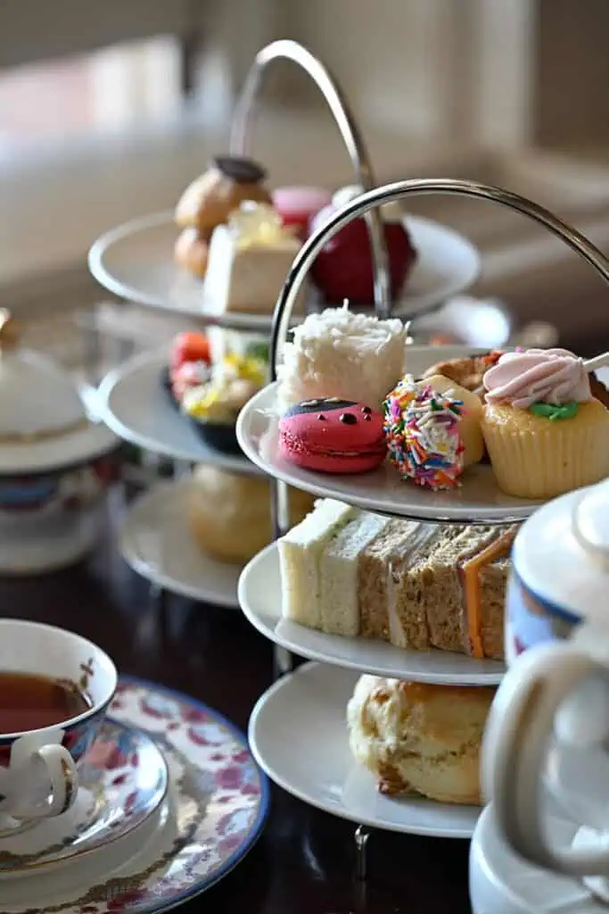 Three-tiered trays with assorted pastries, cupcakes, macarons, finger sandwiches, and scones, alongside a teapot and a floral-patterned teacup with tea on a table.