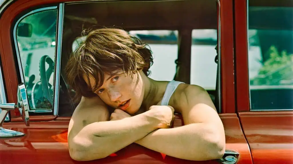 A person with short brown hair leans out of the window of a red vehicle, resting their arms on the window frame and looking directly at the camera.