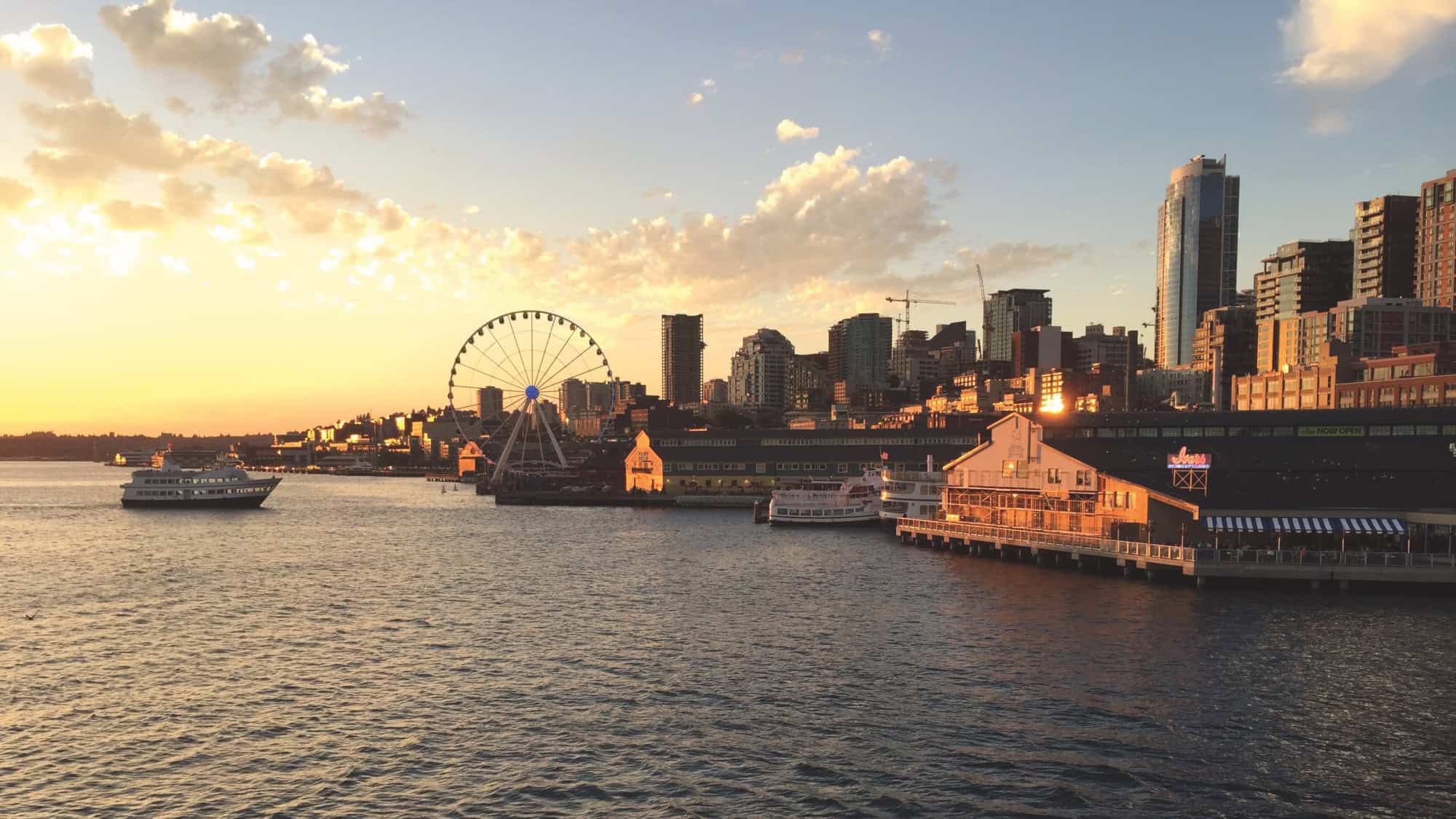 Seattle waterfront at sunset with the Great Wheel, boats on the water, and downtown buildings in the background under a partly cloudy sky—perfect for summer Seattle family outings and stress-free activities.
