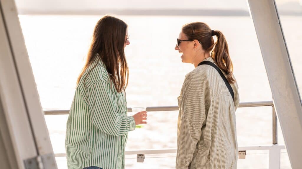 Two women wearing sunglasses and striped shirts stand on a boat deck, talking and smiling, as they enjoy stress-free activities under the summer sun, with sunlight reflecting off the water in the background—a perfect day in the PNW.