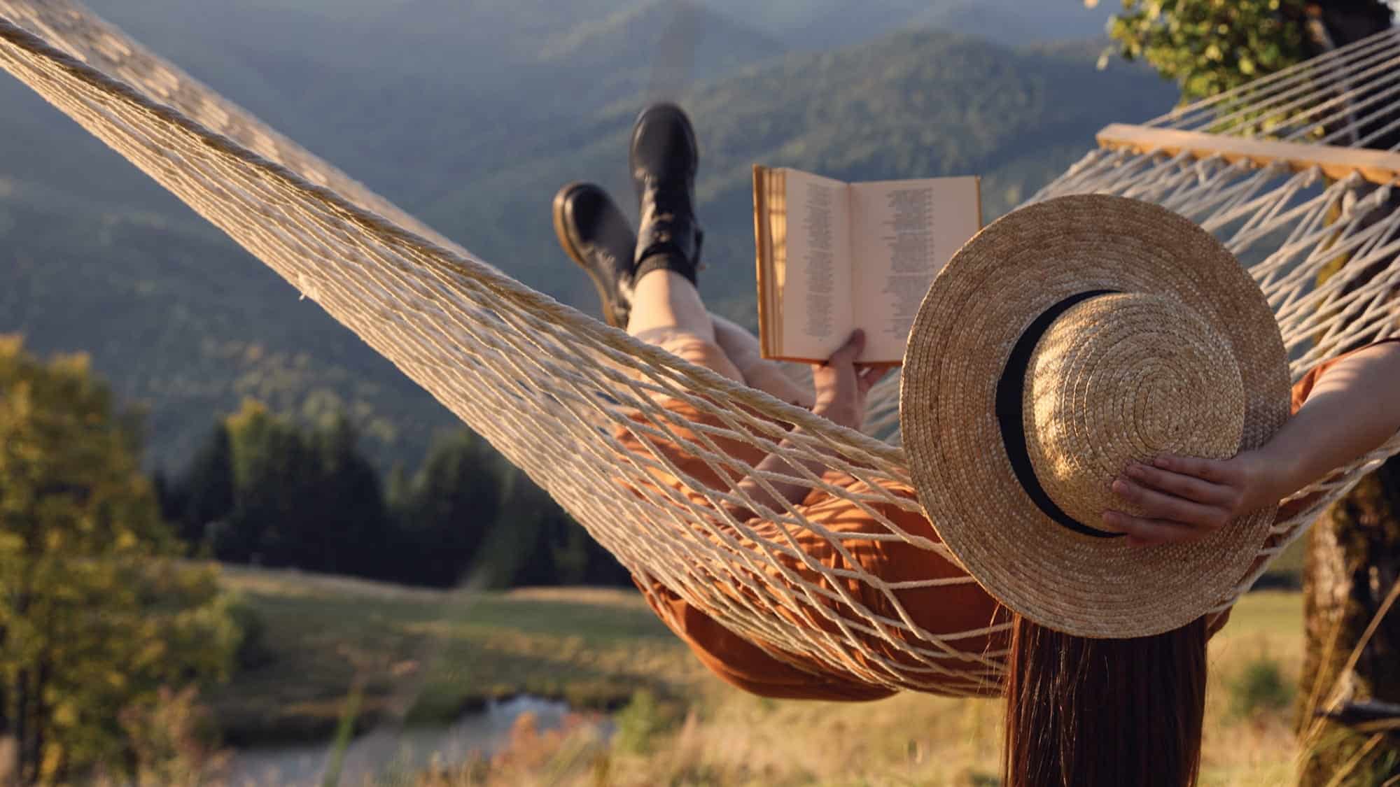 Person wearing a straw hat and boots lies in a hammock outdoors, enjoying a summer read by Washington authors, with mountains and trees visible in the background.