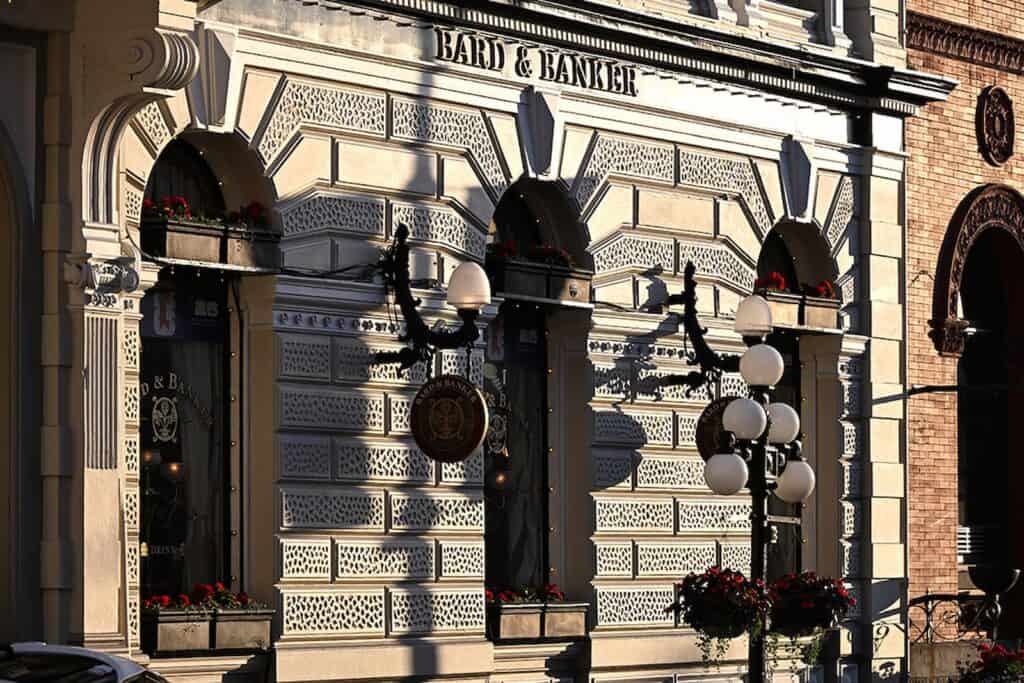 Exterior of the Bard & Banker building with arched windows, ornate facade, hanging signs, and flower boxes in daylight.