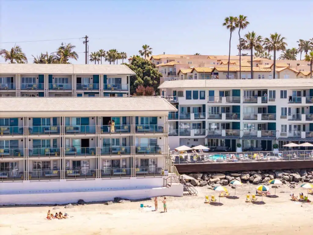 Two multi-story beachfront hotels with balconies face a sandy beach. People are on the sand, some sitting under umbrellas, and palm trees are visible in the background.