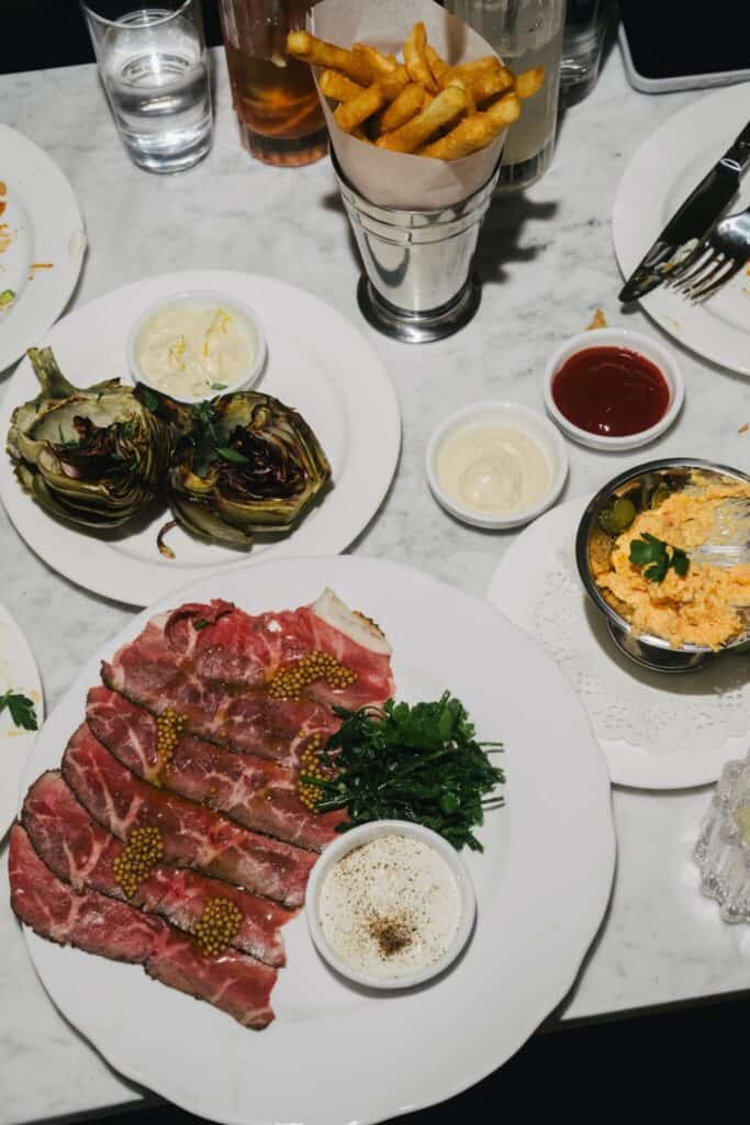 A plate of rare sliced steak with mustard seeds, herbs, and sauce, surrounded by fries, artichokes, and sides on a marble table.