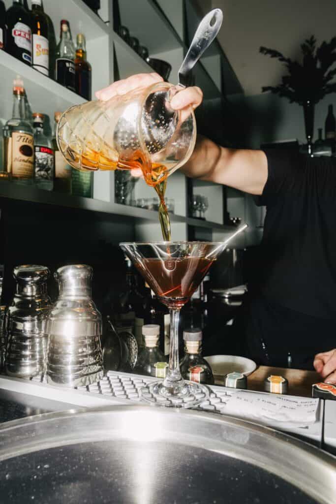 A bartender pours a dark cocktail from a mixing glass into a martini glass at a bar, with liquor bottles and bar tools visible in the background.