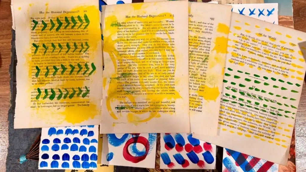 Several old book pages are spread out, each decorated with colorful abstract patterns in paint, including dots, lines, and shapes in yellow, blue, red, and green.