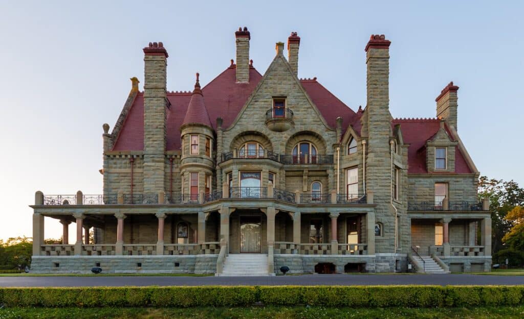 Large historic stone mansion with multiple chimneys, arched windows, wraparound porch, and red roof, viewed from the front against a clear sky—this European-Inspired Getaway offers timeless charm just steps from Seattle’s Backyard.