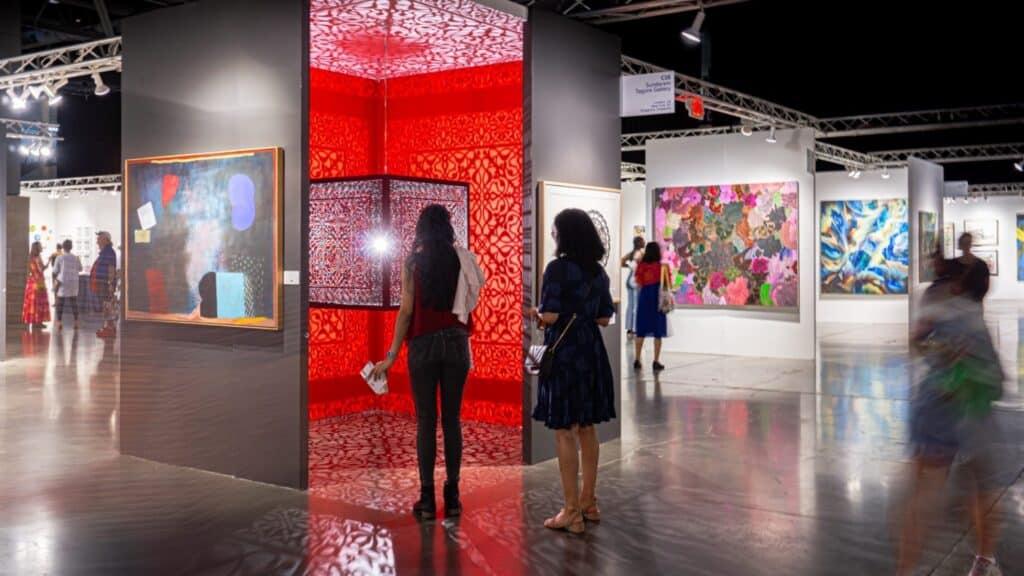 People view colorful paintings and a glowing red art installation at an indoor gallery exhibition.