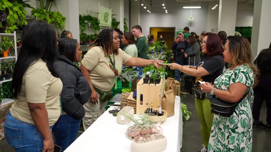 People gather around a vendor table at an indoor plant market, interacting, buying plants, and browsing arrangements in a brightly lit room filled with greenery.