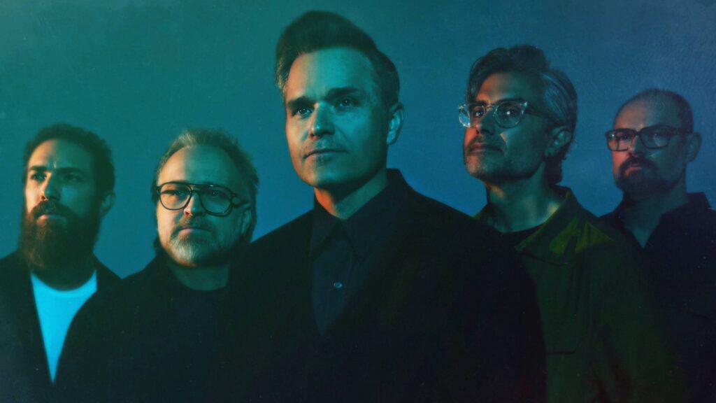 Five men with serious expressions stand together in dim, blue-green lighting, facing slightly to the right, evoking the spirit of Soccer-Themed Art as if ready for a World Cup spotlight.