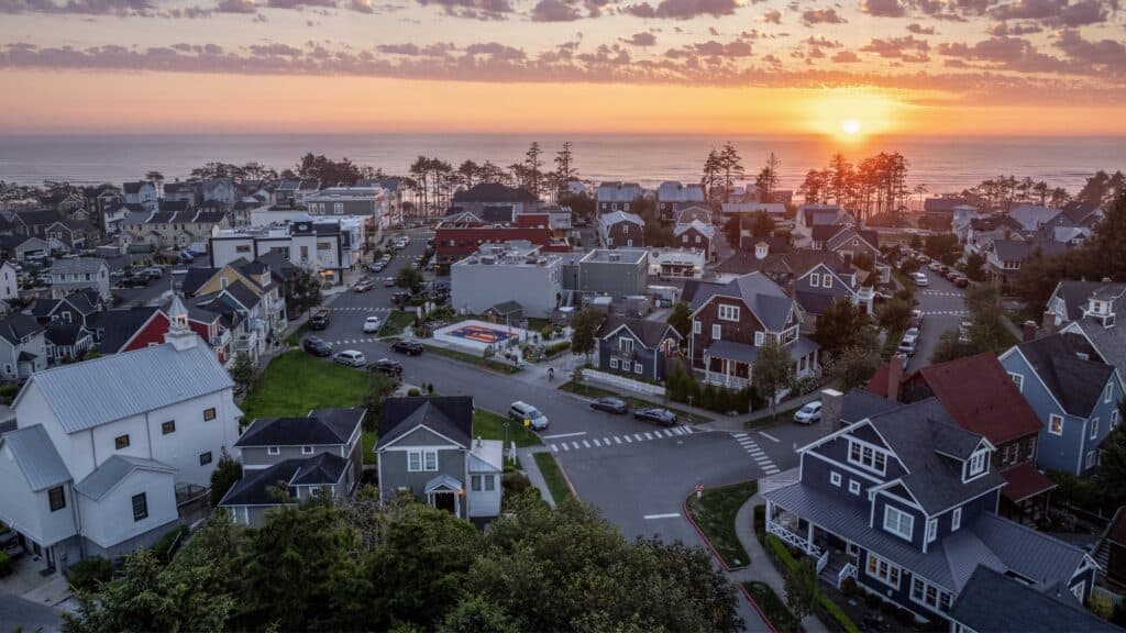 A coastal neighborhood at sunset with neatly arranged houses, intersecting streets, cars parked along the roads, and the sun setting over the ocean in the background.