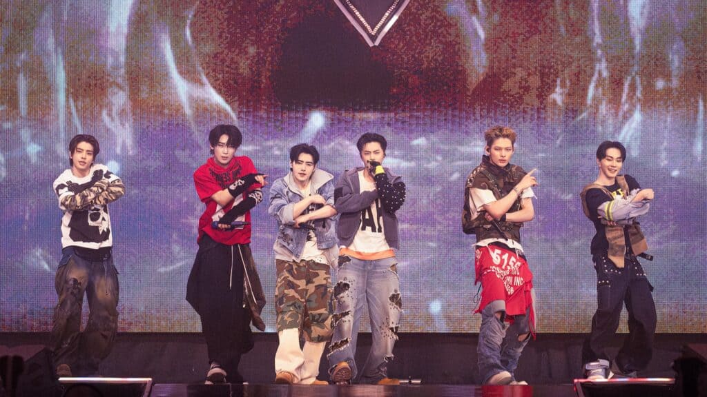 Six male performers stand on stage in coordinated outfits, mid-dance, with crossed arms and a colorful, abstract background behind them.